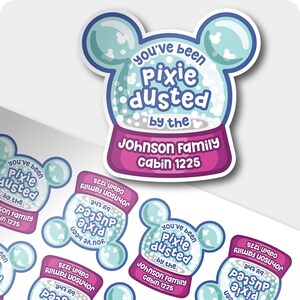 May include: A die-cut sticker shaped like a snow globe with mouse ears, featuring light blue and white colors. The text reads you've been pixie dusted by the Johnson Family Cabin 1225 in a dark blue and white font on a purple base.