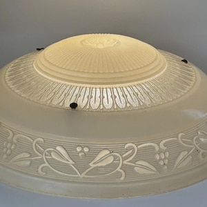 May include: A dome-shaped glass ceiling light shade featuring a frosted finish. The center has a raised sunburst pattern, while the outer rim displays an embossed vine and grape motif. Three small black metal mounting clips are visible on the edge.