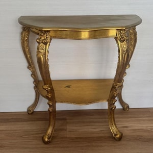 May include: A gold-finished semi-circular console table featuring ornate, carved detailing on the four curved legs and a lower shelf. The table has a smooth, curved top surface and stands on four feet with intricate scrollwork patterns.