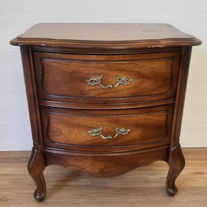 May include: A brown wooden nightstand featuring two curved drawers with ornate brass-toned metal handles. The piece has a smooth top surface and stands on four curved legs, showcasing a rich wood grain finish throughout the structure.