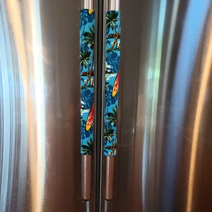 May include: Stainless steel refrigerator with two doors and a bottom freezer drawer. The refrigerator has two handles with blue and white fabric covers featuring a tropical print of palm trees, surfboards, and hibiscus flowers.