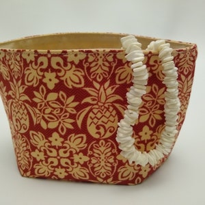 May include: A red and beige patterned tote bag with a white shell necklace draped over the side. The bag features a repeating pineapple and floral design. The interior of the bag is beige.