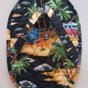 May include: Oval-shaped quilted hot pad with a tropical beach scene. The black fabric features palm trees, vintage cars, and the text "KAUAI, BIG ISLAND, MAUI". It has black handles for easy handling.