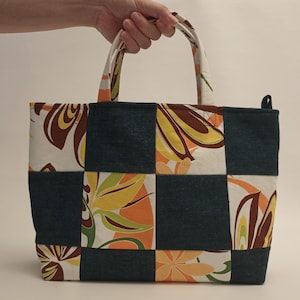 May include: A patchwork tote bag with a floral pattern in orange, yellow, and brown on a white background. The bag has a dark blue denim-like fabric in a checkerboard pattern. The handles are white with a matching floral design.