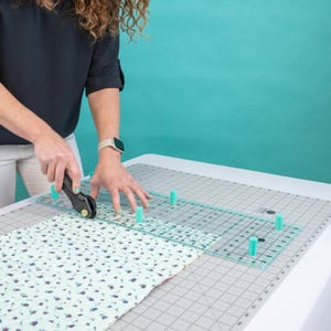 Pode incluir: Uma pessoa usando um cortador rotativo para aparar tecido em uma base de corte. O tecido tem um padrão floral. Uma régua transparente com espaçadores de cor turquesa é usada como guia para o cortador.