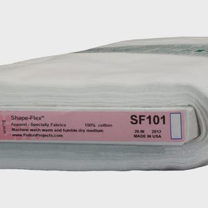 Puede incluir: Un rollo de tela blanca etiquetado como "Shape-Flex SF101" con una etiqueta rosa y blanca. La etiqueta indica que la tela tiene 50.8 cm de ancho y está hecha de 100% algodón. La tela está enrollada y lista para usar.