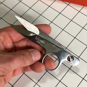 Puede incluir: Tijeras de coser plateadas con punta afilada y un pequeño tornillo en el mango. Las tijeras tienen el texto "V/LDH+ G114" impreso en el mango.