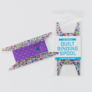 Puede incluir: Una bobina para encuadernación de edredones con un centro de lunares morados y bordes brillantes y coloridos. El embalaje dice "The Original Quilt Binding Spool" en texto azul. Contiene una bobina.