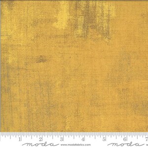 May include: A close-up of a textured fabric with a distressed, mottled appearance. The fabric is primarily a golden yellow colour, with darker, muted brown and grey accents. The fabric has a ruler along the bottom edge.