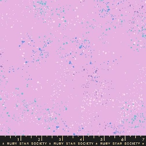 May include: A light purple fabric with a speckled pattern of white, light blue, and dark blue dots. The fabric has a soft, pastel color scheme, and the dots are scattered across the surface. The bottom of the image has a ruler.