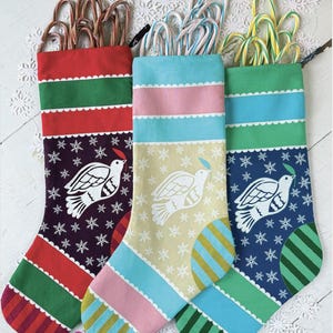May include: Three Christmas stockings filled with candy canes. Each stocking has a different color scheme: red and green, blue and pink, and green and blue. The stockings feature a white dove design and snowflake pattern.