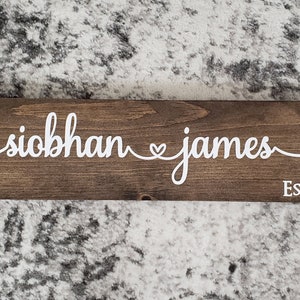 May include: A brown wooden sign with white lettering that reads "siobhan ♥ james EST. 2019".