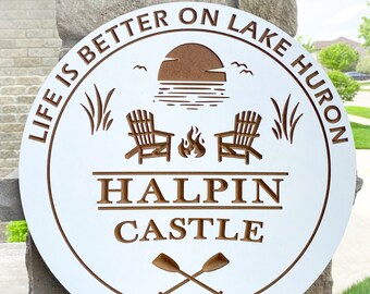 Puede incluir: Señal redonda de madera blanca con texto marrón que dice "Life is better on Lake Huron Halpin Castle Pike Bay, ON". La señal también presenta una silueta marrón de una puesta de sol, dos sillas, una fogata y dos remos.