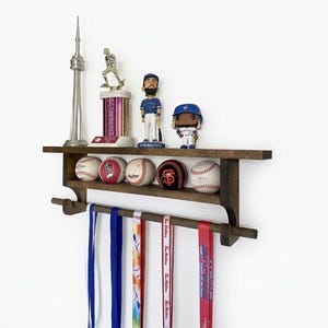May include: A dark brown wooden wall-mounted display shelf featuring a top ledge holding a silver CN Tower replica, a trophy, and two baseball bobbleheads. The middle shelf holds five baseballs, and a bottom rod displays five hanging ribbons.