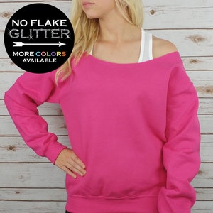 May include: A bright pink off-the-shoulder sweatshirt with long sleeves. The text "NO FLAKE GLITTER" is on a black circle with a white arrow pointing to the right. The text "MORE COLORS AVAILABLE" is below the circle.