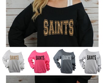 May include: Off-the-shoulder sweatshirt with the word "Saints" in gold glitter lettering. The sweatshirt is available in black, white, pink, and grey.