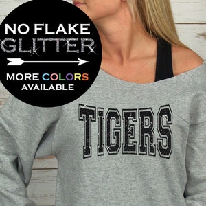 May include: A gray off-the-shoulder sweatshirt with the word "TIGERS" in black, glitter-style lettering across the chest. The sweatshirt has long sleeves and a rounded neckline. A circular graphic in the upper left corner reads "NO FLAKE GLITTER" and "MORE COLORS AVAILABLE."