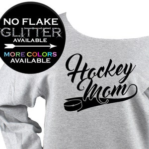 May include: A light gray off-the-shoulder sweatshirt with the words "Hockey Mom" in black script, along with a hockey puck graphic. The image also includes text that says "NO FLAKE GLITTER AVAILABLE" and "MORE COLORS AVAILABLE".