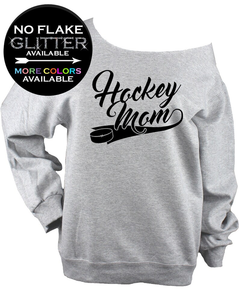 May include: A light gray off-the-shoulder sweatshirt with the words "Hockey Mom" in black script, along with a hockey puck graphic. The image also includes text that says "NO FLAKE GLITTER AVAILABLE" and "MORE COLORS AVAILABLE".