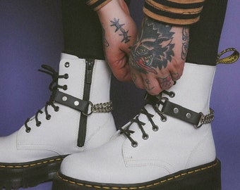 May include: White platform boots with black soles and yellow stitching. The boots have black laces, a side zipper, and a black leather strap with a silver chain detail. The boots are on a purple background.