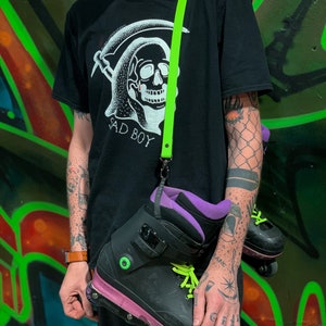 May include: Black inline skates with neon green laces and purple detailing are carried by a neon green strap. The skates are paired with a black t-shirt featuring a skull graphic and the words "SAD BOY".