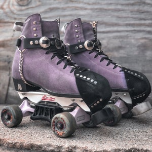 May include: A pair of purple suede roller skates with black toe caps and silver buckles. The skates have black wheels and a white frame. The skates are on a grey concrete surface.