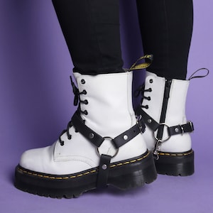 Puede incluir: Botas de cuero blanco con correas y hebillas negras. Las botas tienen una suela negra gruesa y costuras amarillas. Las botas se llevan con pantalones negros.
