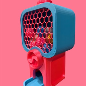 May include: A vibrant gumball machine in red, blue, and pink. The top section is blue with a red honeycomb window. The base is red, and the dispensing area is blue. The machine is filled with assorted coloured gumballs.