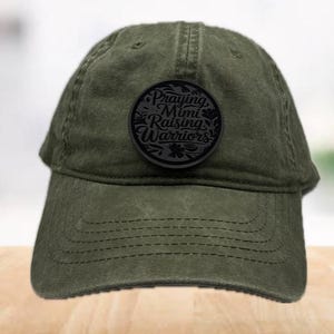 May include: Olive green baseball cap with a curved brim. A black circular patch on the front features the text "Praying Mimi Raising Warriors" in a stylized font, surrounded by floral elements. The cap has a classic, worn look.
