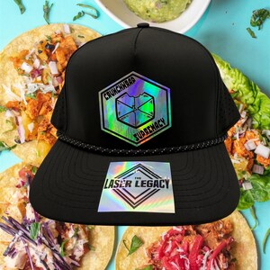 May include: Black trucker hat with a flat peak and a rope detail. The hat features two holographic patches with the text "CRUNCHWRAP SUPREMACY" and "THE LASER LEGACY". The background includes tacos and guacamole.