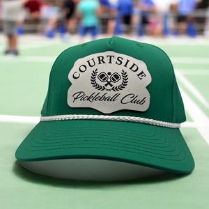 May include: A green Courtside Pickleball Club cap with a white rope detail and a white patch featuring the club's logo. The logo includes crossed pickleball paddles and the text "Courtside Pickleball Club".