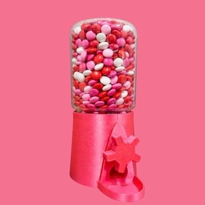 May include: A pink gumball machine with a clear glass jar filled with red, pink, and white candies. The machine has a star-shaped dispensing wheel and a small tray. The background is a solid pink.