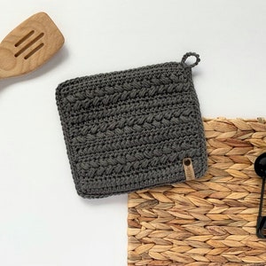 May include: A square, dark gray crocheted potholder with a textured pattern and a small loop for hanging. A small, rectangular tag is attached to the potholder. Wooden kitchen utensils and black measuring spoons are in the background.