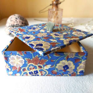 Könnte beinhalten: Eine quadratische Schachtel mit abnehmbarem Deckel, die eine leuchtend blaue Grundfarbe aufweist und mit einem sich wiederholenden Blumenmuster in Creme-, Dunkelblau- und Rotbrauntönen verziert ist. Der Deckel hat eine kleine, salbeigrüne Stofflasche.