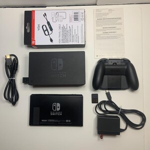 Puede incluir: Un conjunto negro de consola Nintendo Switch y accesorios, dispuestos sobre una superficie blanca. Incluye una base, un mando, un cable HDMI, un adaptador de corriente y una tarjeta de juego. Tambi&eacute;n se ve una caja "YOK HDMI TRAVEL CABLE".