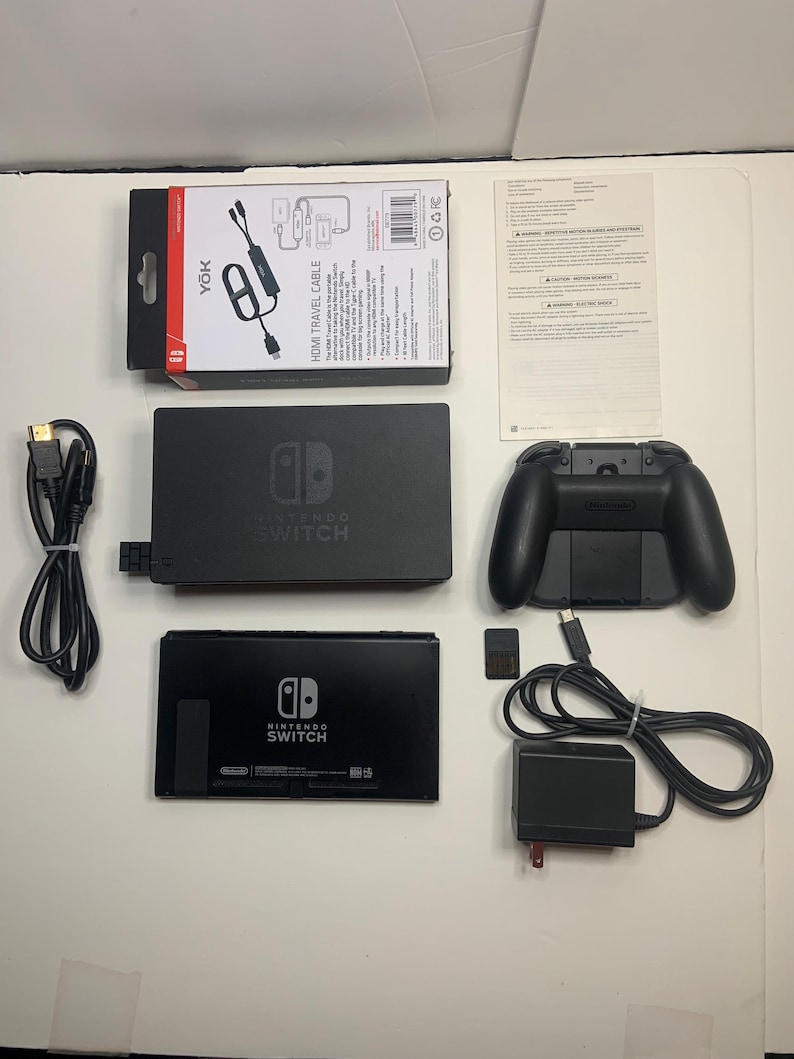 Puede incluir: Un conjunto negro de consola Nintendo Switch y accesorios, dispuestos sobre una superficie blanca. Incluye una base, un mando, un cable HDMI, un adaptador de corriente y una tarjeta de juego. Tambi&eacute;n se ve una caja "YOK HDMI TRAVEL CABLE".