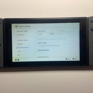 Puede incluir: Una consola Nintendo Switch gris que muestra el men&uacute; de configuraci&oacute;n del sistema. La pantalla muestra opciones para Bluetooth Audio, Bloqueo de pantalla y Gesti&oacute;n de datos. La consola tiene mandos Joy-Con grises adjuntos con botones y joysticks.
