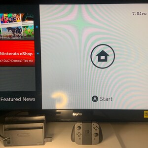 Puede incluir: Un televisor Sanyo negro muestra la pantalla de inicio de Nintendo Switch. La pantalla muestra Nintendo eShop y Noticias destacadas. Un mando y una estaci&oacute;n de acoplamiento Nintendo Switch grises est&aacute;n frente al televisor.