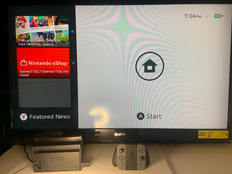 Puede incluir: Un televisor Sanyo negro muestra la pantalla de inicio de Nintendo Switch. La pantalla muestra Nintendo eShop y Noticias destacadas. Un mando y una estaci&oacute;n de acoplamiento Nintendo Switch grises est&aacute;n frente al televisor.