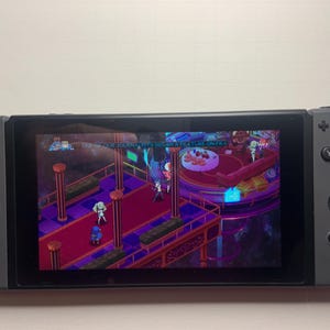 Puede incluir: Una consola Nintendo Switch gris que muestra una escena de videojuego con personajes en una plataforma. El juego presenta una combinaci&oacute;n de colores p&uacute;rpura y rojo con texto en la parte superior. La consola tiene una pantalla negra y mandos grises.