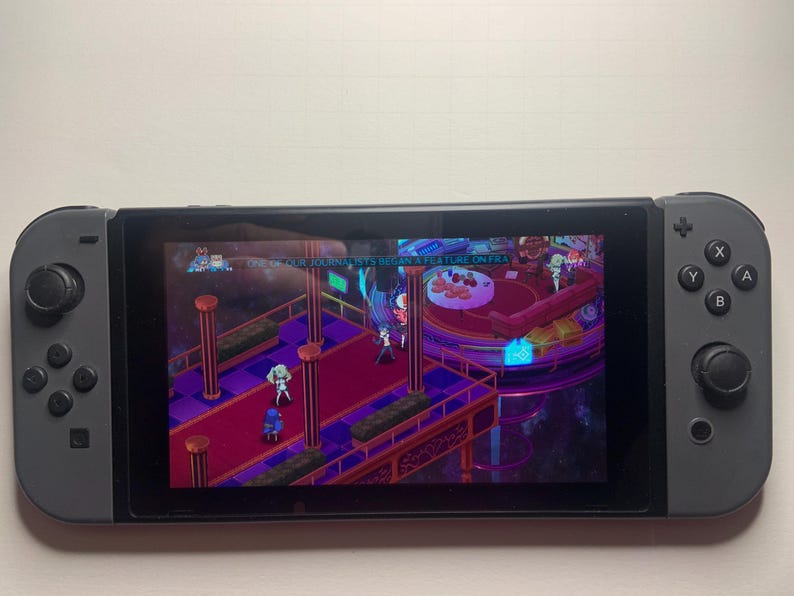 Puede incluir: Una consola Nintendo Switch gris que muestra una escena de videojuego con personajes en una plataforma. El juego presenta una combinaci&oacute;n de colores p&uacute;rpura y rojo con texto en la parte superior. La consola tiene una pantalla negra y mandos grises.