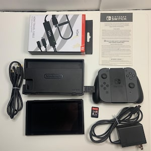 Puede incluir: Un conjunto de consola Nintendo Switch negra, que incluye una base, mandos y pantalla. Los accesorios incluyen un cable de viaje HDMI negro, un adaptador de corriente y una tarjeta de juego. La caja del cable HDMI dice "HDMI TRAVEL CABLE".