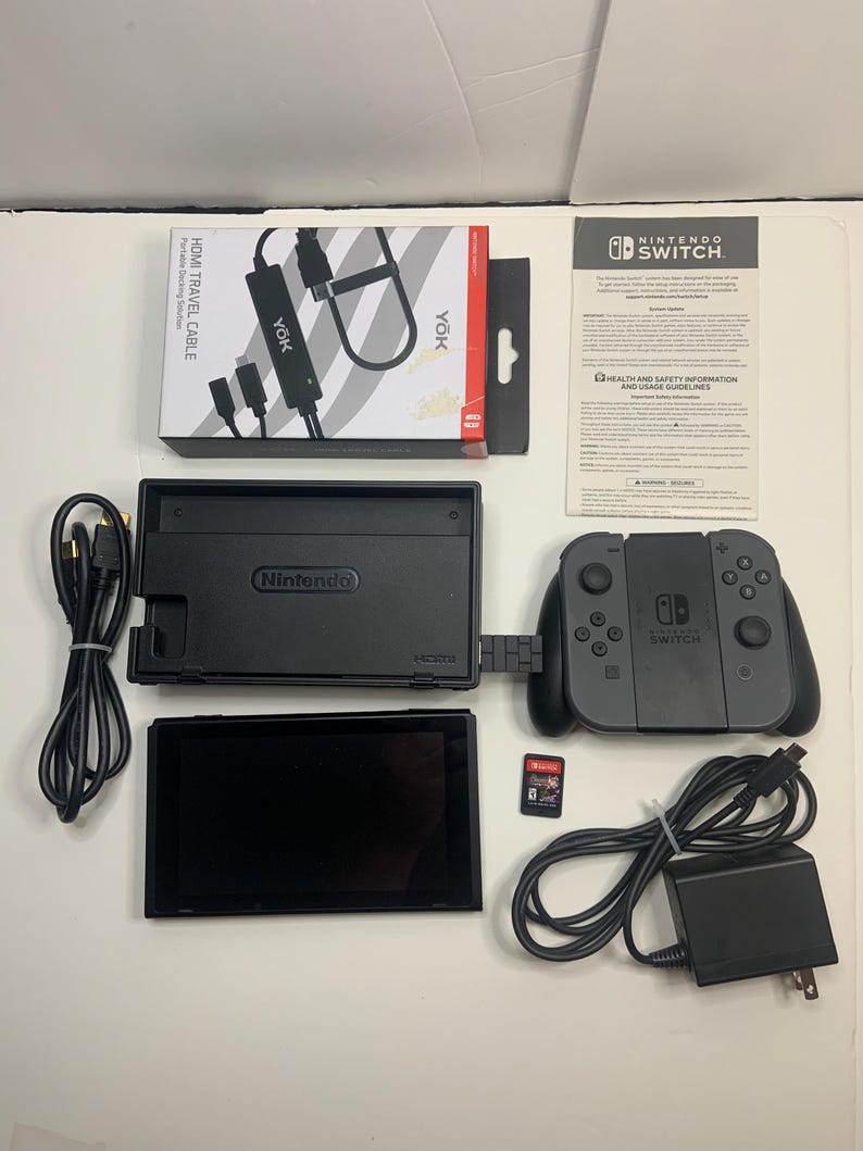 Puede incluir: Un conjunto de consola Nintendo Switch negra, que incluye una base, mandos y pantalla. Los accesorios incluyen un cable de viaje HDMI negro, un adaptador de corriente y una tarjeta de juego. La caja del cable HDMI dice "HDMI TRAVEL CABLE".