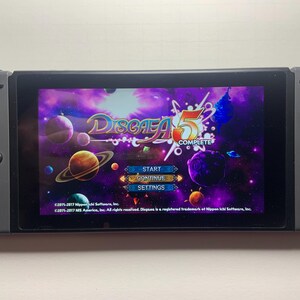 Puede incluir: Una consola Nintendo Switch gris que muestra la pantalla de t&iacute;tulo del juego "Disgaea 5 Complete". La pantalla muestra un fondo espacial p&uacute;rpura y azul con planetas y el logotipo del juego. Los botones y joysticks de la consola son visibles.