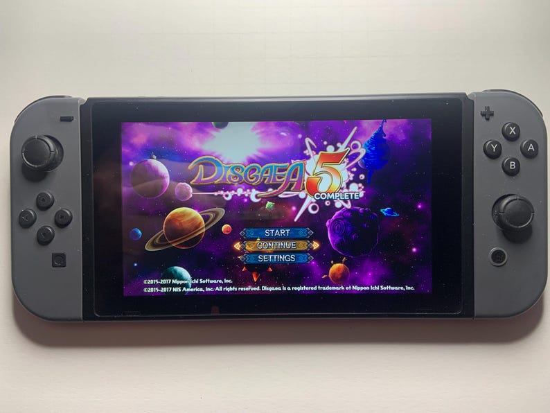 Puede incluir: Una consola Nintendo Switch gris que muestra la pantalla de t&iacute;tulo del juego "Disgaea 5 Complete". La pantalla muestra un fondo espacial p&uacute;rpura y azul con planetas y el logotipo del juego. Los botones y joysticks de la consola son visibles.