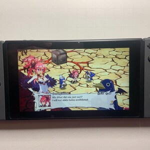 Puede incluir: Una consola Nintendo Switch gris con un juego en la pantalla. El juego presenta personajes de estilo anime y el texto "Seraphina: &iexcl;&iquest;Qu&eacute; acabas de decir!? &iexcl;No tolerar&eacute; ser aniquilada!"