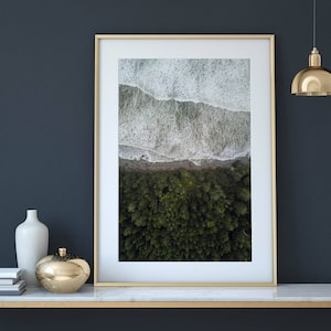 May include: A gold framed picture of an aerial view of a coastline with white waves crashing on a green forest. The picture is hanging on a dark blue wall above a marble console table with gold accents. There are two decorative objects on the table, a white vase and a gold bowl.