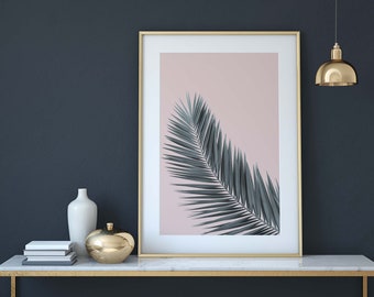 May include: A framed print of a single palm leaf in shades of green against a light pink background. The frame is gold and the print is on a white mat. The print is sitting on a marble console table with a white vase and a gold decorative bowl.