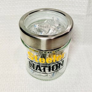 May include: A clear glass jar with a silver lid filled with silver foil-wrapped chocolates. The jar has a black and yellow sticker that says "Steelers Nation".