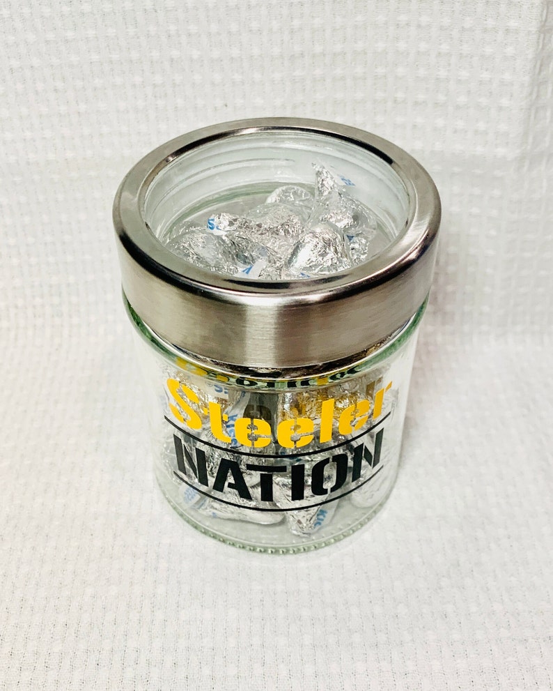 May include: A clear glass jar with a silver lid filled with silver foil-wrapped chocolates. The jar has a black and yellow sticker that says "Steelers Nation".